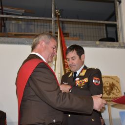 Zwei Männer in Militäruniformen, einer befestigt eine Medaille auf der Brust des anderen in einer formellen Umgebung. Hinter ihnen sind eine Flagge und ein Schild sichtbar.