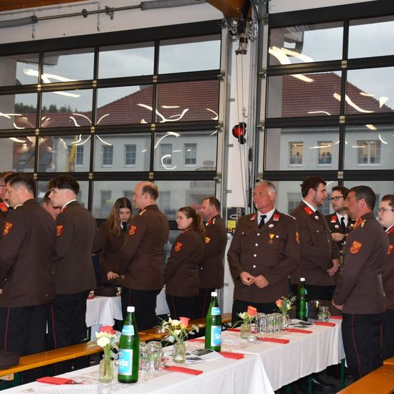 Eine Gruppe uniformierter Personen steht in einem Raum mit großen Fenstern. Sie befinden sich in der Nähe eines Tisches, der mit Blumen und Flaschen geschmückt ist, während eine weitere Gruppe von Personen weiter hinten steht.