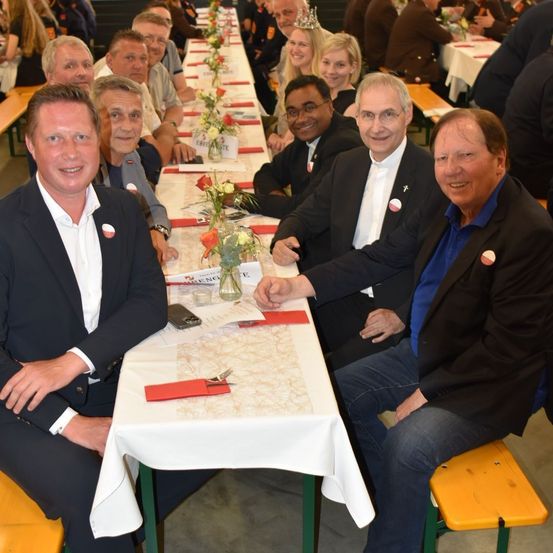 Eine Gruppe von Menschen sitzt an einem langen Tisch in einem Saal. Sie sind formell gekleidet. Der Tisch ist mit einer weißen Tischdecke bedeckt. Es gibt Blumen und einen Namensschild auf dem Tisch. Sie lächeln und posieren für ein Foto.