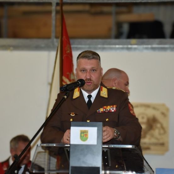Ein Mann in Uniform steht an einem Podium und spricht in ein Mikrofon. Hinter ihm weht eine Fahne, und ein weiterer Mann steht in der Nähe. Im Hintergrund befinden sich weitere Personen.
