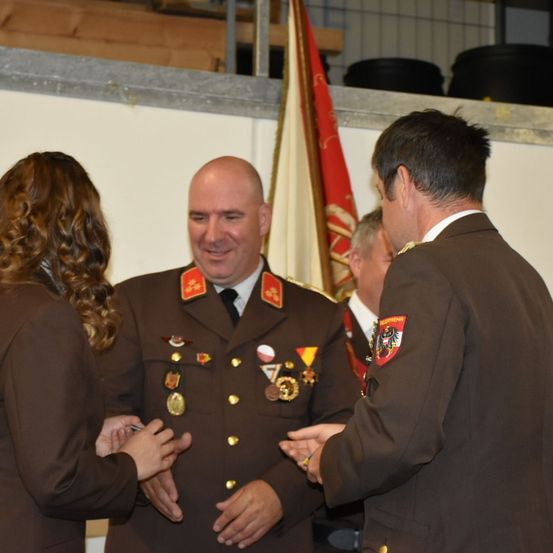 Ein Mann in Uniform schüttelt die Hände einer Frau und eines anderen Mannes, vor einer Flagge stehend.