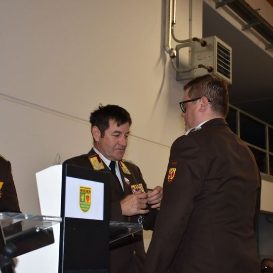 Zwei Militäroffiziere stehen vor einem Podium, einer scheint dem anderen eine Auszeichnung zu überreichen. Der Offizier, der die Auszeichnung erhält, hält eine Medaille.