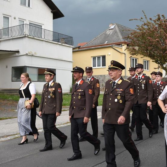 Eine Gruppe von Militäroffizieren in braunen Uniformen marschiert die Straße entlang. Hinter ihnen gehen zwei Frauen, eine hält eine Handtasche. Gebäude säumen die Straße, eines mit einem Balkon und ein anderes mit einem Baum davor.