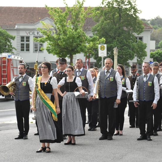 Eine Gruppe von Musikern und Beamten in traditioneller Kleidung, mit Instrumenten und Bannern, steht in einer Parade auf einer Straße mit einem Gebäude und Bäumen im Hintergrund.