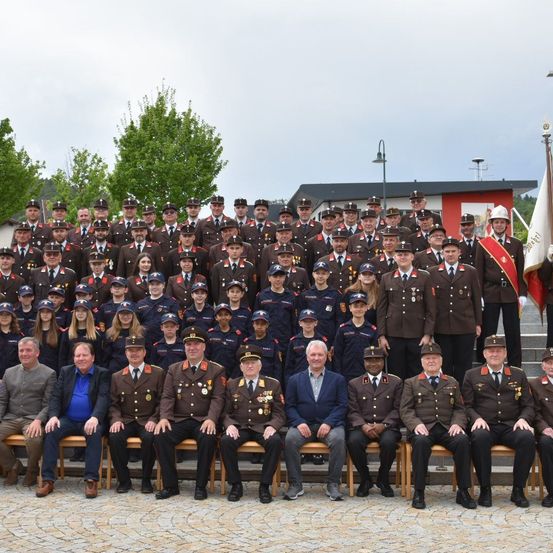 Eine große Gruppe uniformierter Personen, darunter Offiziere und Kadetten, ist in einer Außenumgebung versammelt, wobei einige auf Bänken sitzen. Sie sind von Bäumen und Gebäuden umgeben.