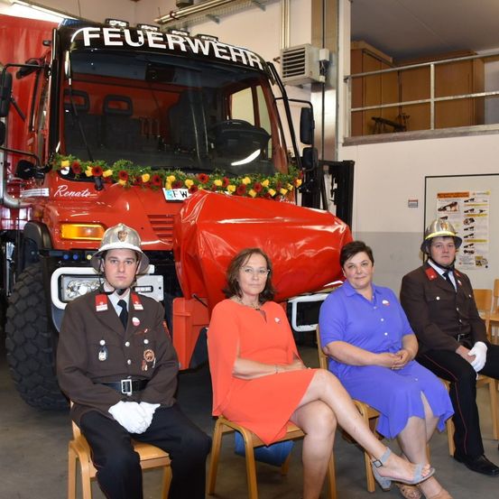 Vier Personen in Uniformen und Mützen sitzen auf Stühlen vor einem roten Feuerwehrauto. Hinter ihnen steht eine weiße Wand mit einem Schild. Ein Feuerwehrauto mit Blumen auf der Front ist vor ihnen geparkt.