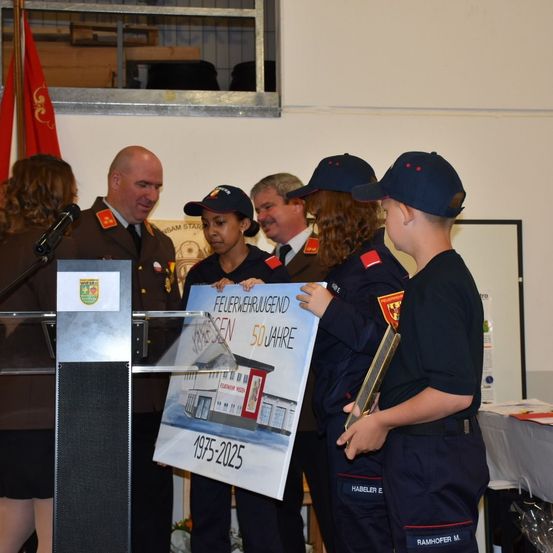 Eine Gruppe von Menschen steht um ein Podium herum mit einem Schild, auf dem 'Feuerwehrjugend 50 Jahre 1975-2025' steht. Einige von ihnen halten Auszeichnungen. Hinter ihnen steht ein Mann in Uniform und eine Frau mit einem Mikrofon. Links ist eine Flagge und eine Holzkiste. Dahinter befindet sich ein Glasfenster.