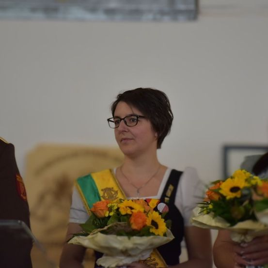 Eine Frau mit kurzen Haaren, die eine Brille und eine Schärpe trägt, hält einen Blumenstrauß. Sie steht in einem Raum mit weißen Wänden und einer großen Steinstruktur im Hintergrund.