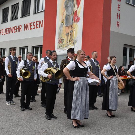 Eine Gruppe von Menschen steht vor einem Gebäude, einige halten Musikinstrumente, andere sind in traditioneller Kleidung gekleidet, möglicherweise während einer Veranstaltung.