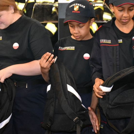 Drei Feuerwehrleute helfen einem Kind dabei, einen Rucksack anzuziehen. Das Kind trägt einen Hut mit der Aufschrift 'Feuerwehr'. Dahinter stehen zwei weitere Feuerwehrleute.