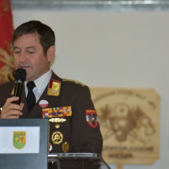 Ein Mann in Uniform spricht in ein Mikrofon an einem Podium mit einem Logo. Dahinter ist ein verschwommenes Bild einer Flagge und eines hölzernen Schildes.