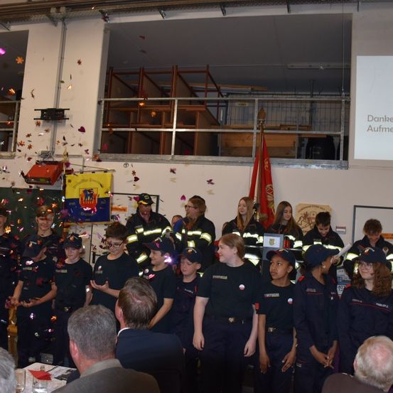 Eine Gruppe von Kindern in Uniform steht vor einer Menge, einige mit Mützen und Brillen. Ein Flagge und ein Banner sind im Hintergrund zu sehen. Konfetti fällt.
