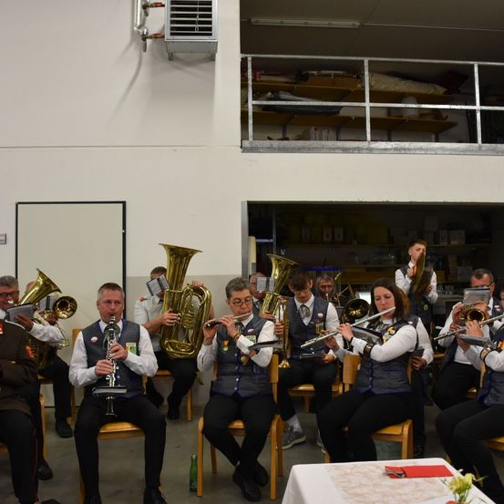 Eine Gruppe von Musikern in Uniform sitzt auf Stühlen und spielt Blechblasinstrumente in einem Raum mit weißer Wand und Holzboden. Ein Balkon und Regale sind im Hintergrund sichtbar.