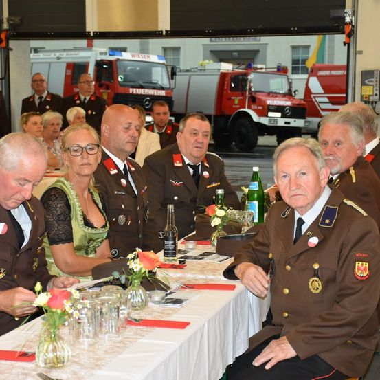Eine Gruppe formell gekleideter Personen, möglicherweise Feuerwehrleute, sitzen an einem Tisch mit Blumen und Flaschen. Dahinter sind mehrere Feuerwehrfahrzeuge geparkt.