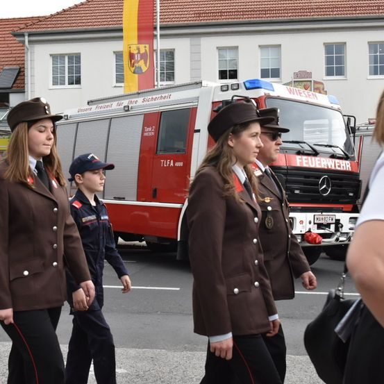 Drei junge Frauen und ein Junge in Uniform gehen an einem Feuerwehrauto vorbei. Sie sind auf der Straße, und hinter ihnen steht ein weißes Gebäude mit Fenstern und einem Fahnenmast.