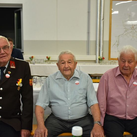 Drei ältere Männer sitzen auf einer Bank. Der Mann auf der linken Seite hat eine Medaille an seiner Uniform. Der Mann in der Mitte trägt ein gestreiftes Hemd. Der Mann auf der rechten Seite trägt ein rosa Hemd. Hinter ihnen ist ein Tisch mit Blumen und Tassen aufgebaut.