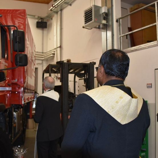 Zwei Männer stehen in einem Lagerhaus, einer trägt eine Brille und eine goldene Stola, der andere einen schwarzen Anzug. Im Hintergrund sind ein Gabelstapler und ein roter Lkw zu sehen.