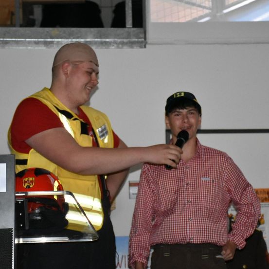 Ein Mann mit einem Verband auf dem Kopf hält ein Mikrofon an einen anderen Mann in einem roten karierten Hemd, der in die Kamera lächelt.