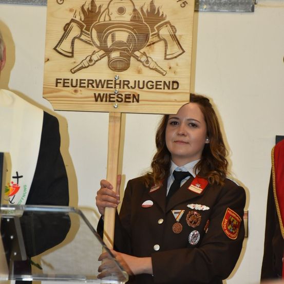 Eine Frau in Uniform hält ein Holzschild mit dem Text 'Feuerwehrjugend Wiesen'. Sie steht vor einem Podium mit einem Mann in einem schwarz-weißen Gewand hinter ihr.