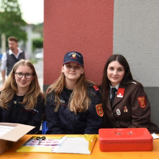 Drei Frauen in Uniform stehen neben einem Tisch mit Gegenständen darauf und lächeln für ein Foto. Eine rote Box und eine Schere liegen auf dem Tisch. Sie tragen Mützen und Uniformen mit Abzeichen. Ein Mann in einer Weste steht hinter ihnen.