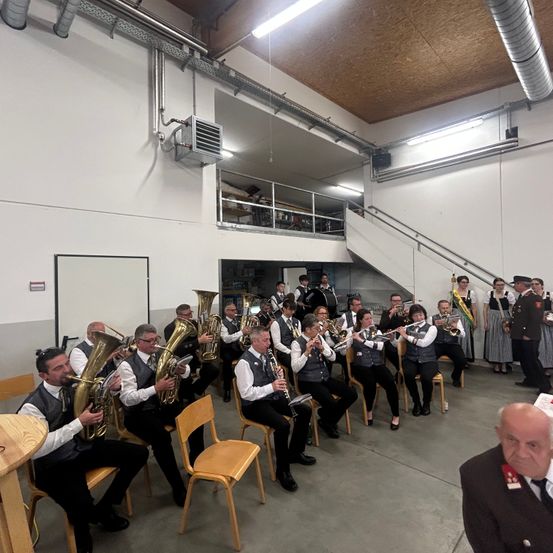 Eine Band spielt in einem großen Raum mit Stühlen und Tischen. Sie spielen Instrumente wie Tuben, Trompeten und Klarinetten. Einige sitzen, andere stehen. Im Hintergrund befinden sich eine Treppe und ein Balkon.