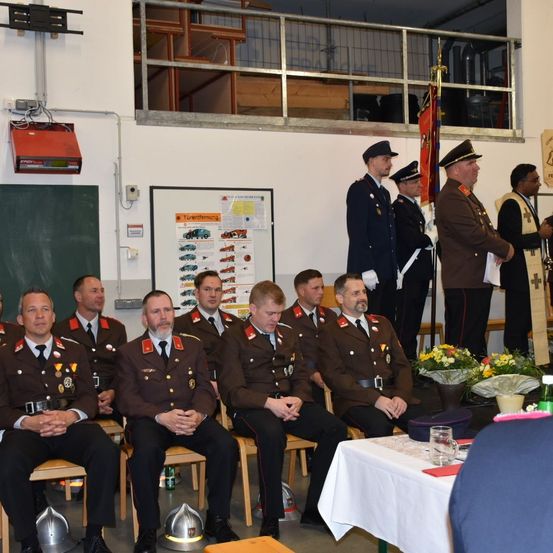 Eine Gruppe uniformierter Personen sitzt auf Stühlen, möglicherweise während einer Zeremonie. Sie werden von anderen in Uniform begleitet, die hinter ihnen stehen, mit einer Flagge und einem Banner im Blick. Ein Tisch mit Blumen und einer Flasche ist im Vordergrund.