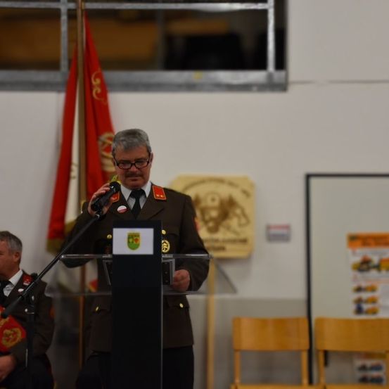 Ein Mann in Uniform spricht in ein Mikrofon an einem Podium mit Fahnen dahinter. Ein leeres Podium und Stühle sind davor.