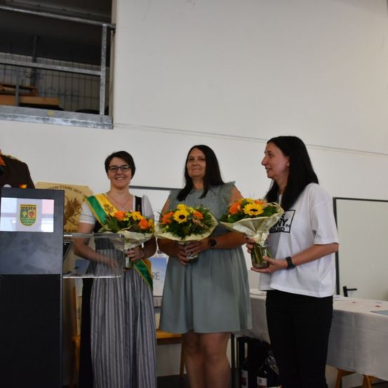 Drei Frauen stehen in einem Raum und halten Blumensträuße in den Händen, eine lächelt und schaut die anderen an. Ein Podium und ein Tisch mit einer weißen Tischdecke sind in der Nähe.