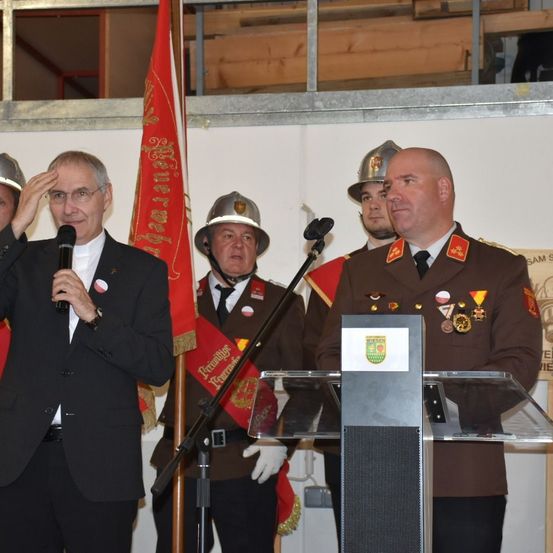 Ein Mann mit Brille hält ein Mikrofon und salutiert. Hinter ihm stehen zwei Männer in Militäruniformen mit einer Fahne. Ein kahlköpfiger Mann in Uniform steht neben einem Podium.