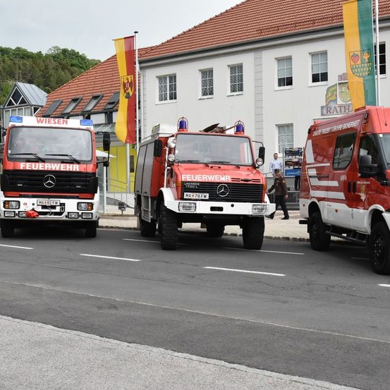 Drei Feuerwehrfahrzeuge sind auf der Straße vor einem Gebäude mit rotem Dach geparkt. Zwei Fahnen befinden sich an der Seite des Gebäudes. Eine Person geht auf dem Bürgersteig.
