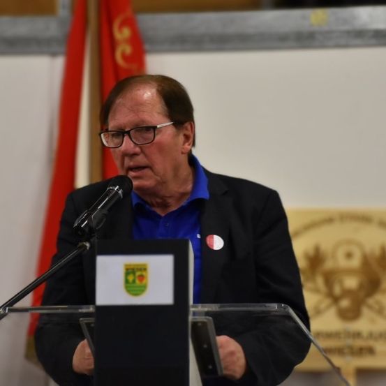 Ein älterer Mann mit Brille und blauem Hemd hält eine Rede an einem Rednerpult mit Mikrofon. Dahinter ist eine rote Fahne zu sehen.