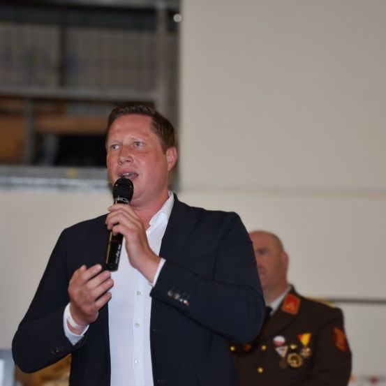 Ein Mann in einem dunklen Anzug spricht in ein Mikrofon, möglicherweise bei einer formellen Veranstaltung. Hinter ihm steht ein Offizier in Uniform.