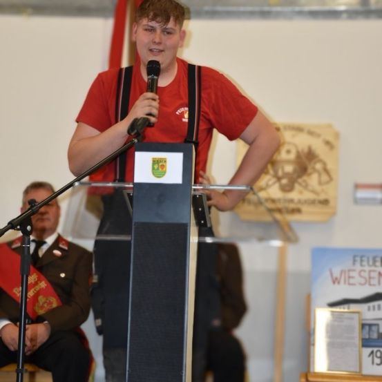 Ein junger Mann steht an einem Rednerpult und spricht in ein Mikrofon. Er trägt ein rotes Shirt und eine Hose mit Trägern. Hinter ihm hört ein Mann in Militäruniform zu. Die Umgebung umfasst eine weiße Wand mit verschiedenen Bannern und einer verschwommenen Figur.