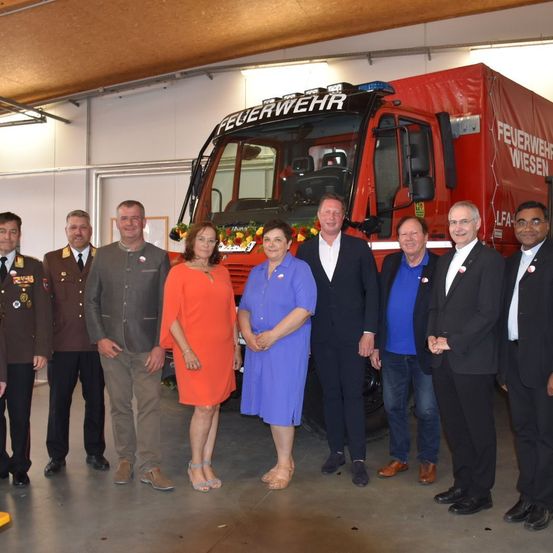 Eine Gruppe von Menschen steht vor einem roten Feuerwehrauto mit dem Wort Feuerwehr darauf. Eine Frau trägt ein orangefarbenes Kleid, und eine andere Frau trägt ein blaues Kleid. Hinter ihnen tragen einige Männer Anzüge.