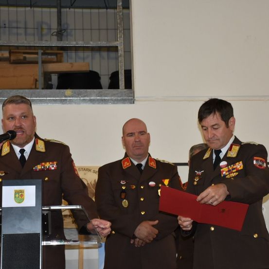 Drei Männer in Militäruniformen stehen in einem Raum. Einer liest aus einer roten Mappe, während die anderen zuhören. Ein Podium mit Mikrofon steht vor dem Mann auf der linken Seite. Hinter ihnen befindet sich ein Metallgeländer mit Lagerboxen.