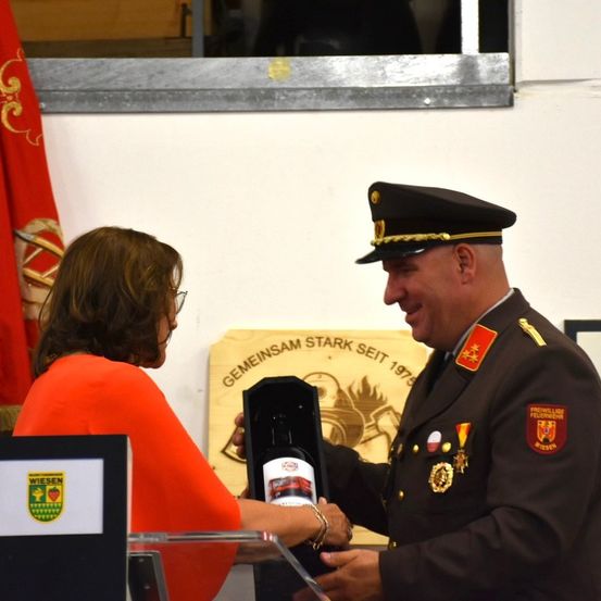Eine Frau und ein Mann in Uniform tauschen eine Flasche in einer Box während einer Veranstaltung aus. Die Frau trägt eine Brille und der Mann Medaillen. Ein Fahnen und ein Schild sind im Hintergrund.