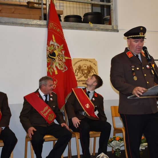 Bei einer formellen Veranstaltung sind mehrere Personen in Militäruniformen zu sehen, von denen eine an einem Podium eine Rede hält. Hinter ihnen ist eine Fahne mit einem Wappen sichtbar.