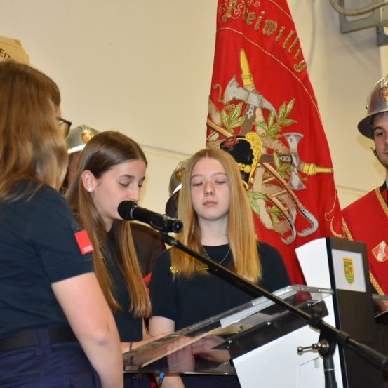 Ein Mädchen spricht in ein Mikrofon an einem Rednerpult mit einer Flagge dahinter, während ein anderes Mädchen zusieht. Ein drittes Mädchen steht neben ihnen.