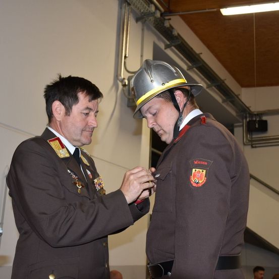 Ein uniformierter Mann steckt einem anderen uniformierten Mann, der einen Helm trägt, in einem Raum eine Abzeichen an.