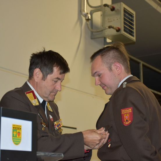 Zwei Männer in Militäruniformen sind drinnen. Einer befestigt eine Plakette am anderen. Sie stehen vor einem weißen Schild mit einem Logo und einem Logo an der Wand hinter ihnen.