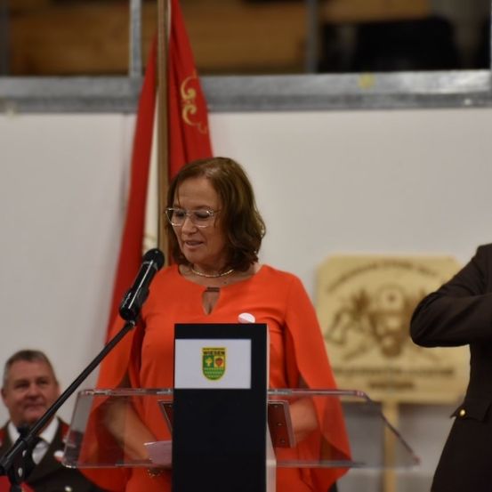 Eine Frau in einem orangefarbenen Kleid spricht an einem Rednerpult mit Mikrofon, wobei eine Fahne und eine weitere Person hinter ihr stehen.