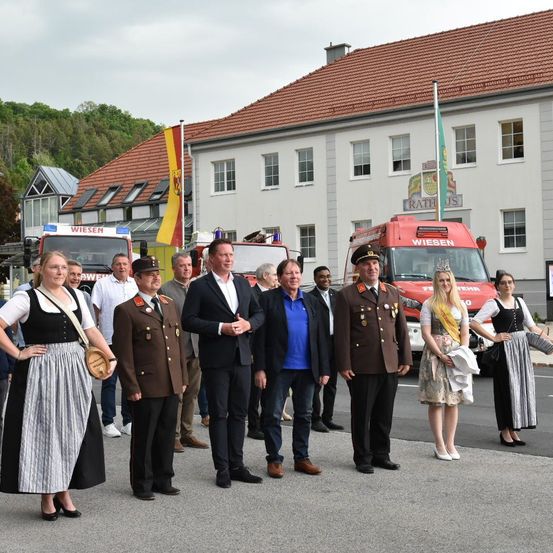 Eine Gruppe von Menschen, darunter Feuerwehrleute und ein Militäroffizier, steht vor einem weißen Gebäude mit einem roten Dach. Das Gebäude hat Fenster und eine Fahne auf einem Mast. Hinter ihnen sind Feuerwehrwagen geparkt. Eine Frau in einer traditionellen Tracht steht links und eine Frau mit einer Krone rechts.
