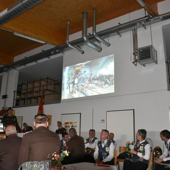 Eine Gruppe von Menschen in formeller Kleidung sitzt in einem Raum. Eine Leinwand projiziert ein Bild, und einige Personen stehen. Ein Tisch mit Gläsern und Blumen steht davor.