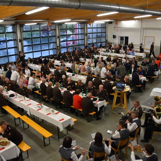 Eine große Indoor-Veranstaltung mit vielen Menschen, die an Tischen sitzen. Musiker spielen im Vordergrund Instrumente. Die Tische sind mit weißen Tischdecken bedeckt, einige haben Blumen und Flaschen. Der Raum hat große Fenster, und Autos sind draußen sichtbar.