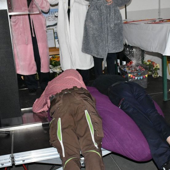 Drei Personen sind anwesend, wobei zwei in der Nähe eines Podiums stehen und eine dritte auf einem violetten Kissen liegt. Dahinter steht ein mit einem weißen Tuch bedeckter Tisch.