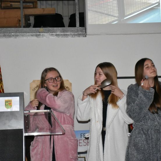 Drei Frauen stehen in einem Raum. Die Frau auf der linken Seite lächelt und hält ein Mikrofon. Die Frau in der Mitte hält einen Löffel. Die Frau auf der rechten Seite hält eine Zigarette. Hinter ihnen sind ein Podium und eine Wand mit einem Poster.