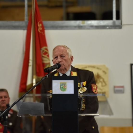 Ein älterer Mann in Uniform steht an einem Rednerpult und spricht in ein Mikrofon. Hinter ihm ist eine rote Fahne.