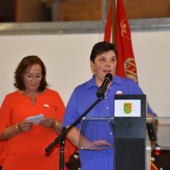 Eine Frau in einem blauen Shirt spricht in ein Mikrofon an einem Rednerpult. Eine andere Frau in einem orangefarbenen Shirt steht hinter ihr und hält ein Stück Papier. Hinter ihnen ist eine rote Fahne zu sehen.