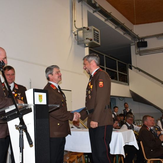 Zwei Männer in Militäruniformen schütteln sich die Hände vor einem Podium. Sie sind von mehreren Personen umgeben, die an einem Tisch sitzen. Die Umgebung scheint eine formelle Veranstaltung oder Zeremonie zu sein.