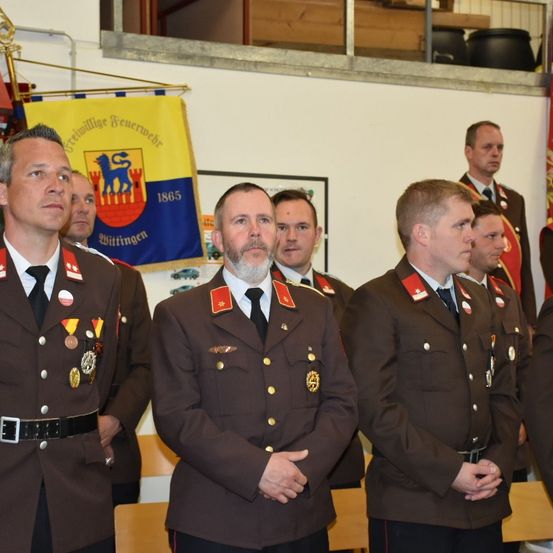 Eine Gruppe uniformierter Personen, möglicherweise Feuerwehrleute, steht vor einem Banner und einer Galerie mit Fahnen.